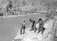 CCC RIVER TRAIL CONSTRUCTION: STAGED PHOTO WITH FOREMAN WITH PIPE IN MOUTH POINTING TO TRAIL WORK. 2 ENROLLEES WORKING. BLACK BRIDGE OVER COLORADO RIVER BEYOND. CIRCA 1934 STAFF. 