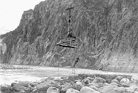 DETAIL OF CCC ENROLLEES SUSPENDED ABOVE THE COLORADO RIVER IN A CABLE TRAMWAY CAR. MAN HANGING BELOW THE TRAM CAR FROM A ROPE LADDER. CIRCA 1936. NPS.